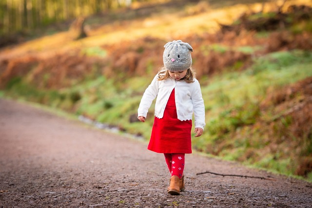 A little girl in a red dress and hat walking on a dirt road